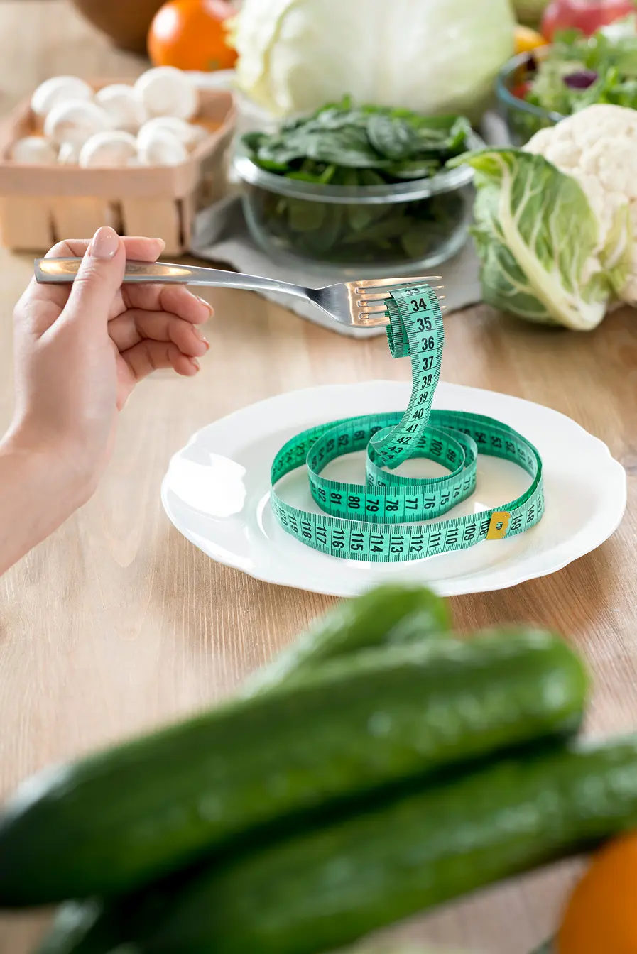 A person holding a fork with measuring tape on a white plate surrounded by fresh vegetables.