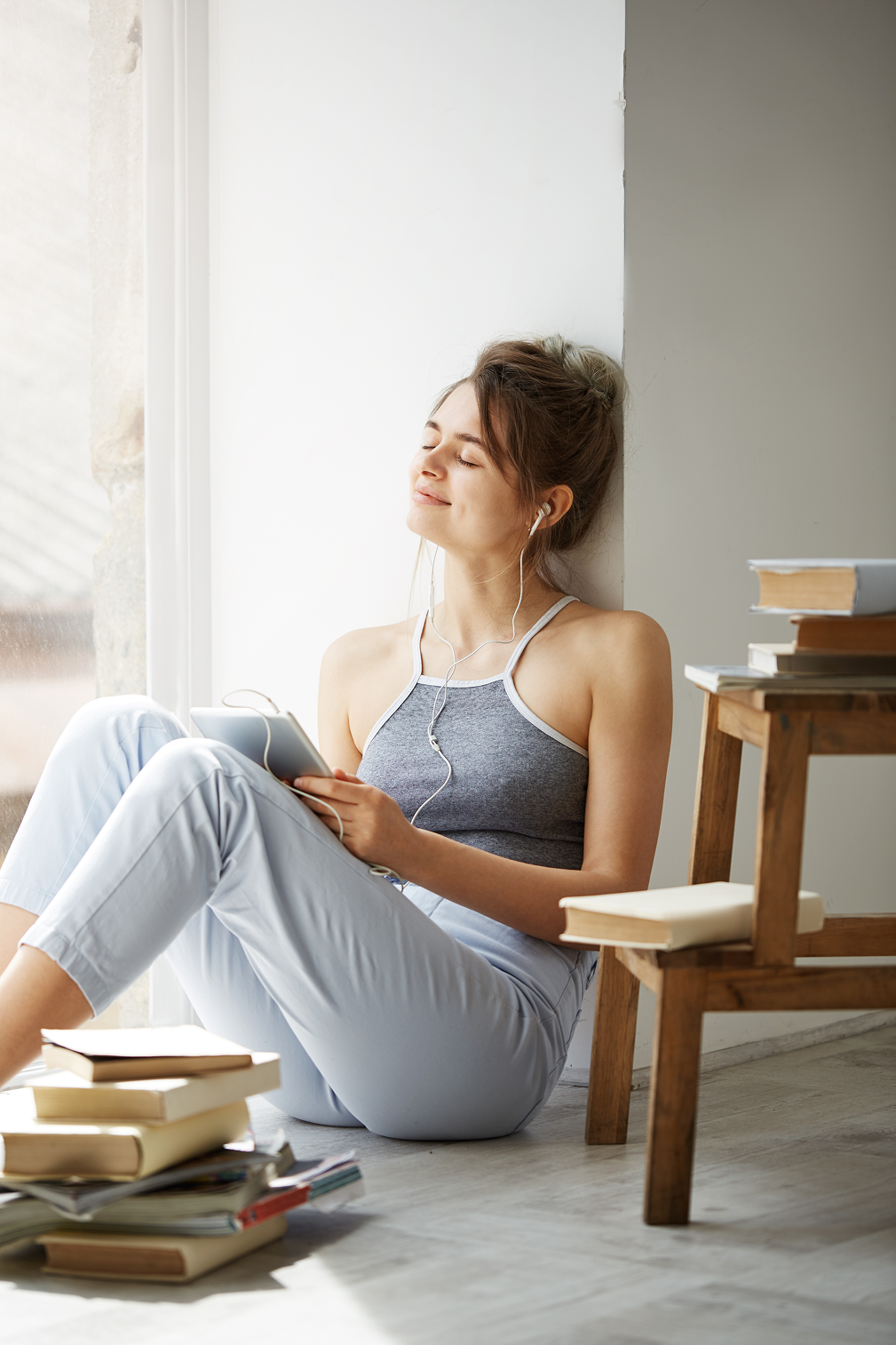 A woman smiling with closed eyes, listening to music on her tablet with headphones, surrounded by books.
